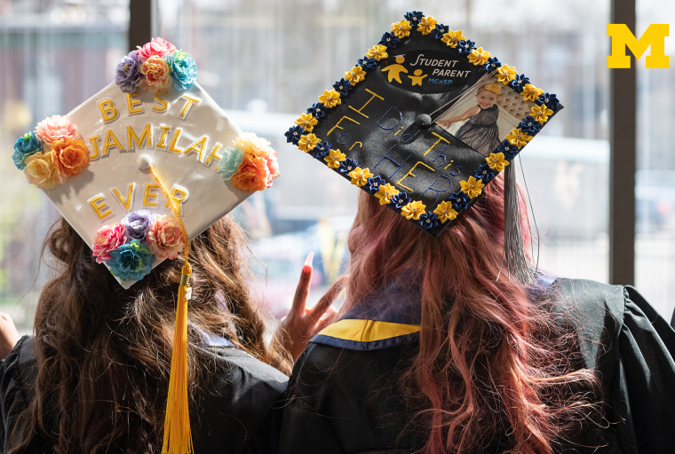 2 graduates with backs to the camera wearing decorated graduation caps and giving the peace sign