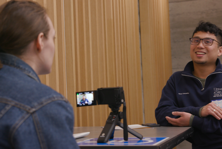 Two students sit across from each other, engaged in conversation, while one films the interview