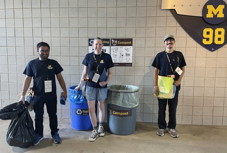 Three stadium volunteers stand in front of bin signage.