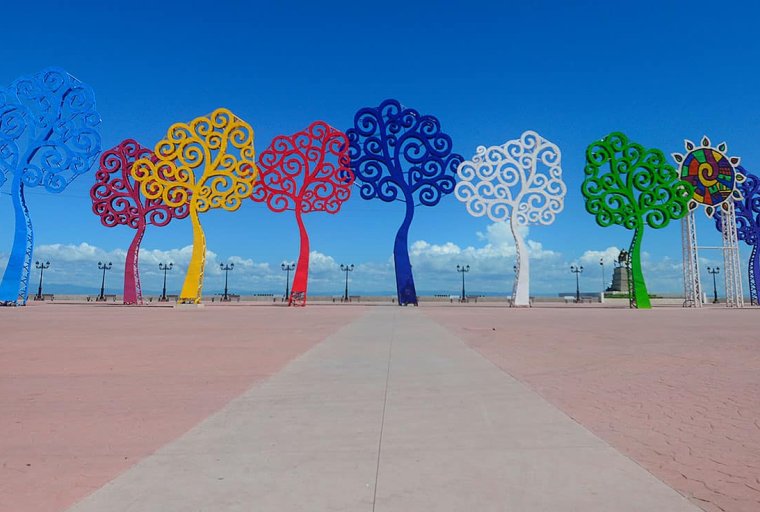 Vibrant metal tree sculptures in red, yellow, blue, and green line a pink walkway under a clear blue sky.