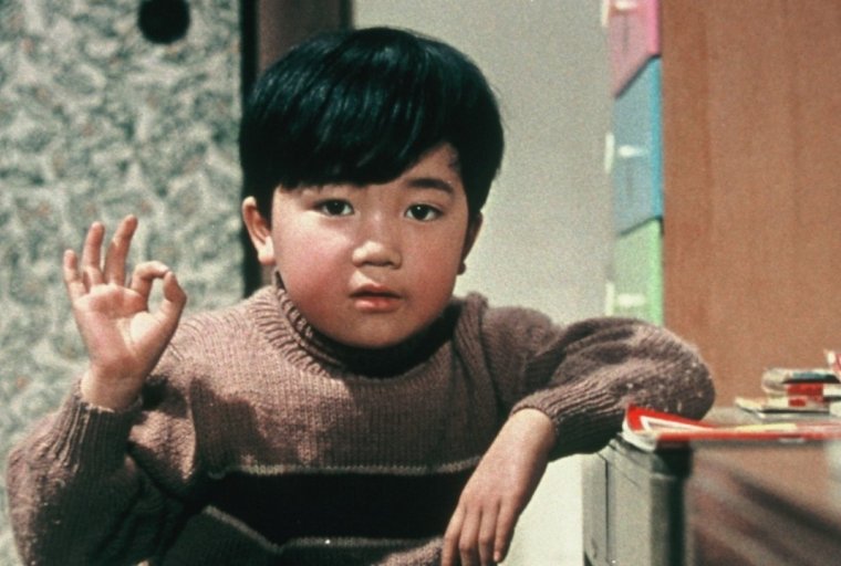 A film still of a young Japanese boy making the "ok" sign in a 1950s-style room