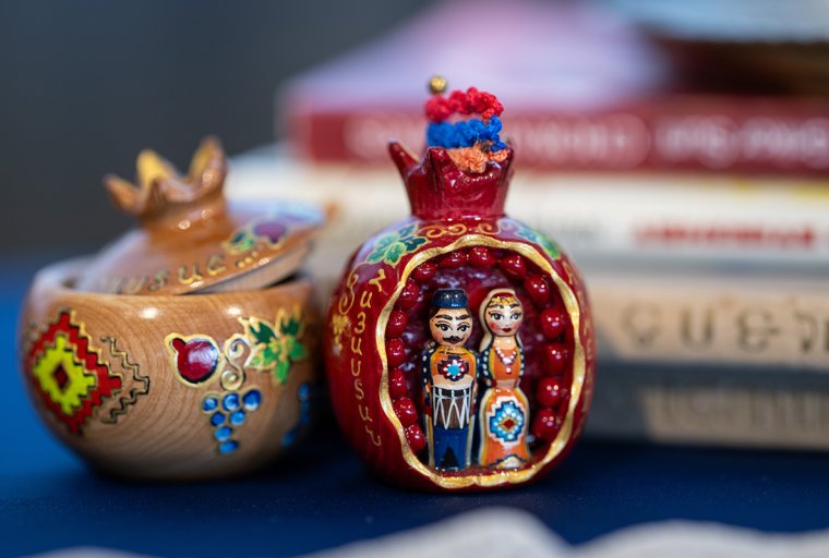 Close-up photograph of two small decorative Armenian objects displayed on a table. On the right is a red, pomegranate-shaped ornament opened to reveal painted figurines of a man and a woman wearing traditional Armenian clothing, framed by small red bead details. On the left is a light wooden pomegranate-shaped container with a removable lid, decorated with hand-painted Armenian motifs in red, green, blue, and yellow. Blurred books are visible in the background, emphasizing the objects as cultural artifacts associated with memory and tradition.