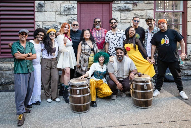 Photo of 14 member interracial troupe posed against an outdoor wall space, with two large drums in foreground.
