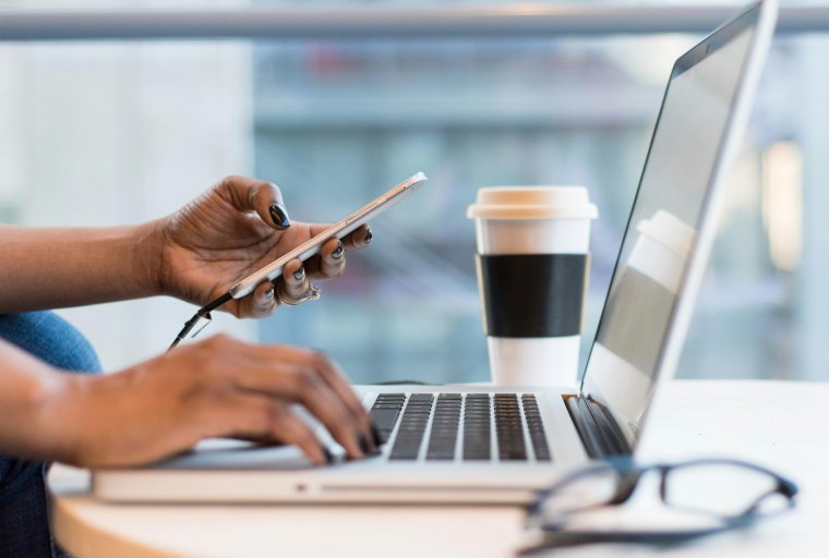 Image of person sitting at a short table holding their phone and working on their laptop, with a coffee nearby.