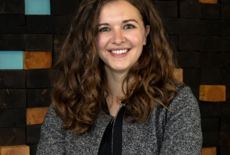 Allison Winstel, a woman with long, wavy brown hair, smiles at the camera with her arms crossed. She is wearing a gray jacket over a dark top and stands in front of a textured wall made of stacked wooden blocks in dark and natural tones.