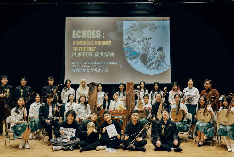 A large group photo of members of the Qingyun Chinese Music Ensemble on a concert stage. Around twenty performers are arranged in several rows, some seated and some standing, holding traditional Chinese musical instruments such as pipa, erhu, guzheng, ruan, and bamboo flutes. A projection screen behind them displays the concert title along with the ensemble’s name. The performers wear coordinated black, white, and green concert attire. The stage has wooden flooring and theatrical lighting, creating a formal concert setting.