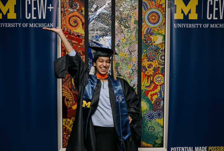 A joyful graduate in University of Michigan cap and gown poses with one arm raised, set against a vibrant mosaic backdrop. Two university banners flank her.