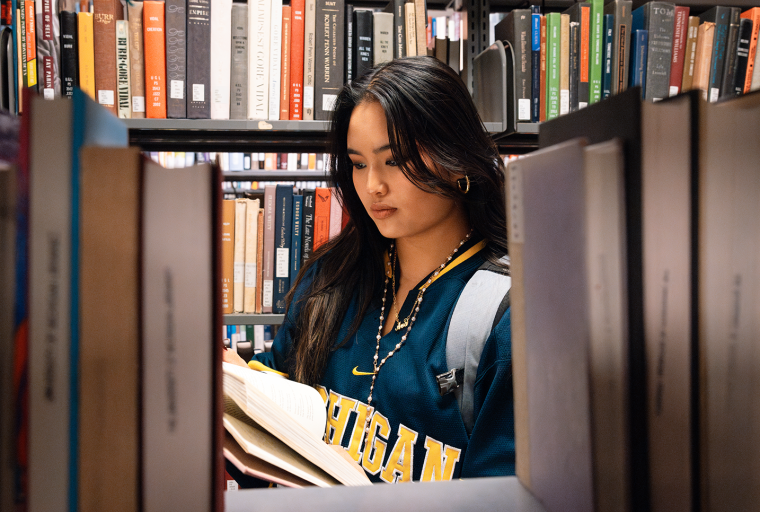 A woman wearing a Michigan sweatshirt reads a book in a library aisle, surrounded by shelves filled with books.