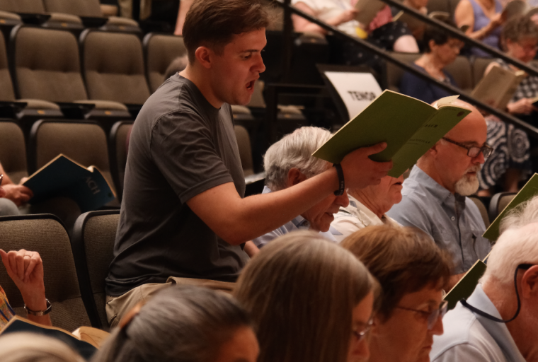 Male singer stands in tiered seating holding music binder.