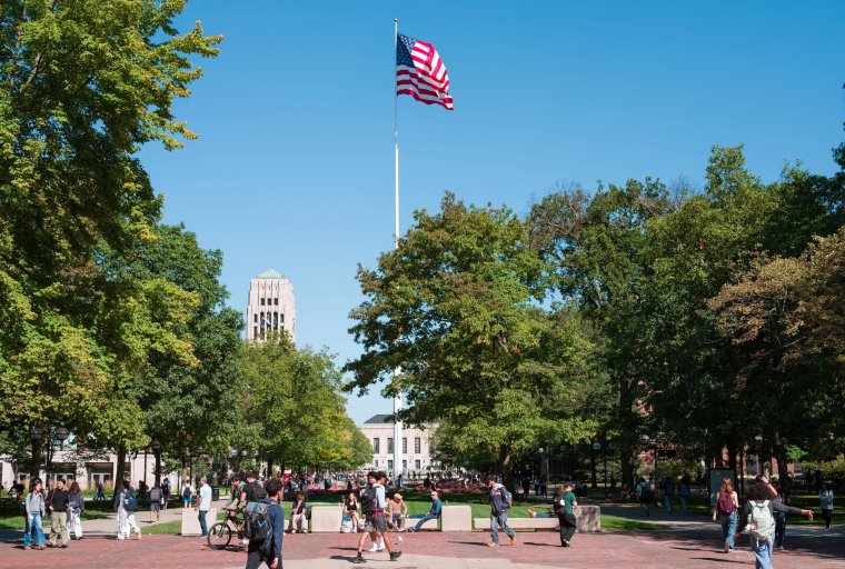 People walking across the Diag with an American flag and Burton Tower in the background, surrounded by green trees under a clear blue sky.
