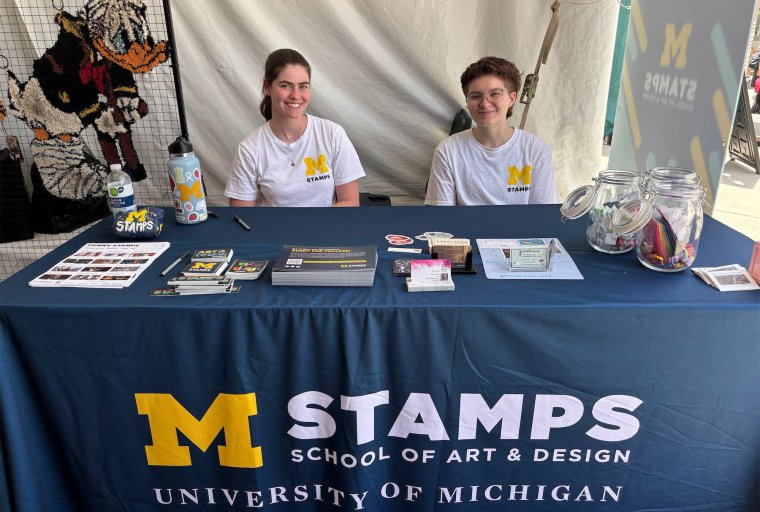 Two students, smiling, sit at a table covered with a blue table adorned with an M STAMPS logo
