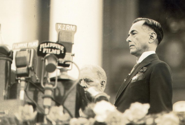 Vintage photo of a man in a suit (Manuel Quezon) standing near a bank of microphones at an outdoor speech.