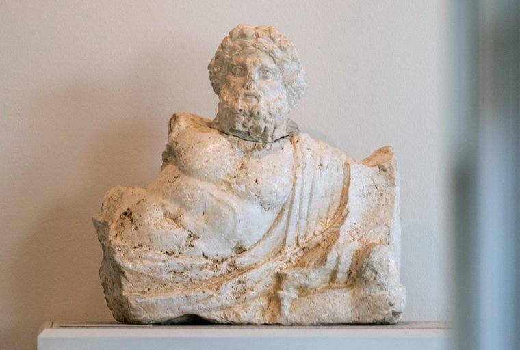 White travertine sculpture of the torso and head of Nilus, a bearded and curly-haired male figure in a semi-reclined pose.