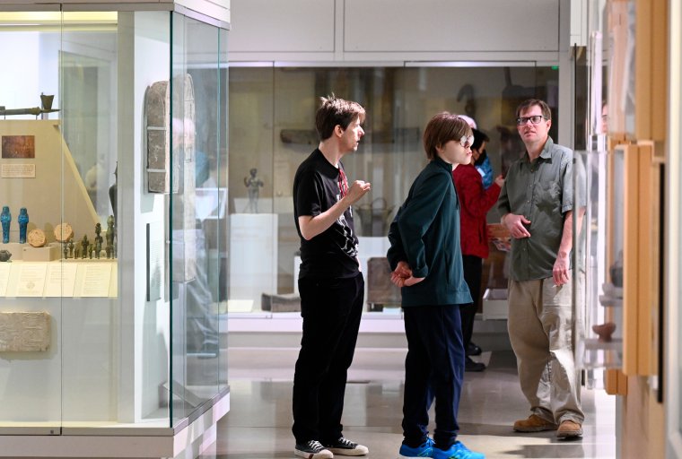 A family of visitors pauses between the Dynastic and Graeco-Roman Egyptian galleries at the Kelsey Museum. Displayed in a case to their left are several small Egyptian artifacts, including bronze figurines of deities and faience ushabtis.