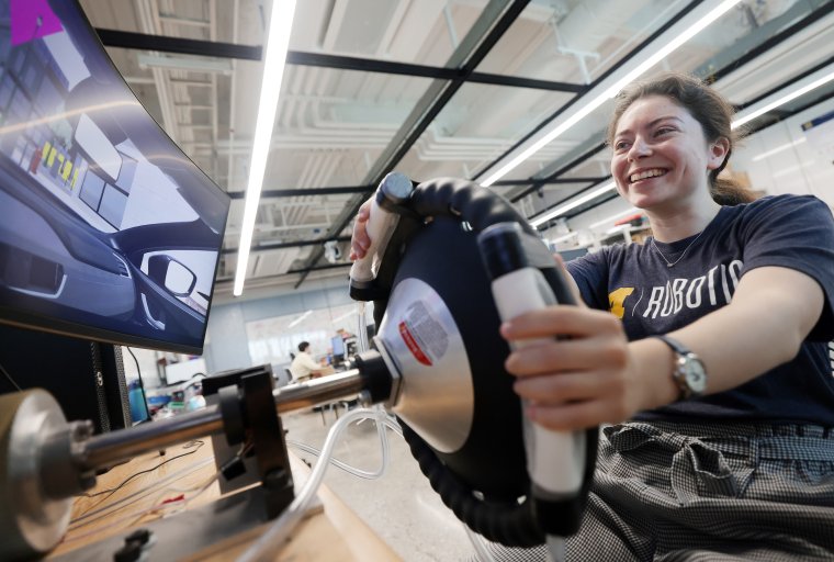 Hannah tests out a steering wheel with hands on its haptic feedback sensors while driving in a lab looking at the monitor.