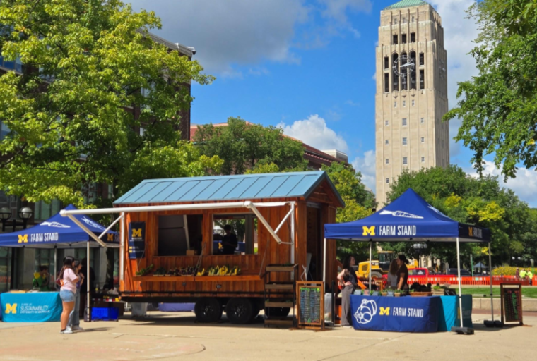 A wooden farm stand trailer sits in front of the belltower on S Ingalls mall with shelves filled with local produce and two tents on either side