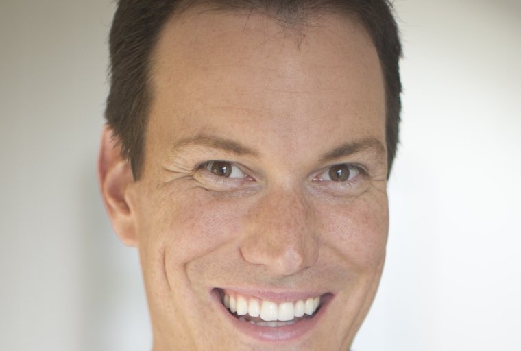 Headshot of Shawn Achor smiling, wearing a dark suit jacket over a light-colored collared shirt against a soft, out-of-focus background.