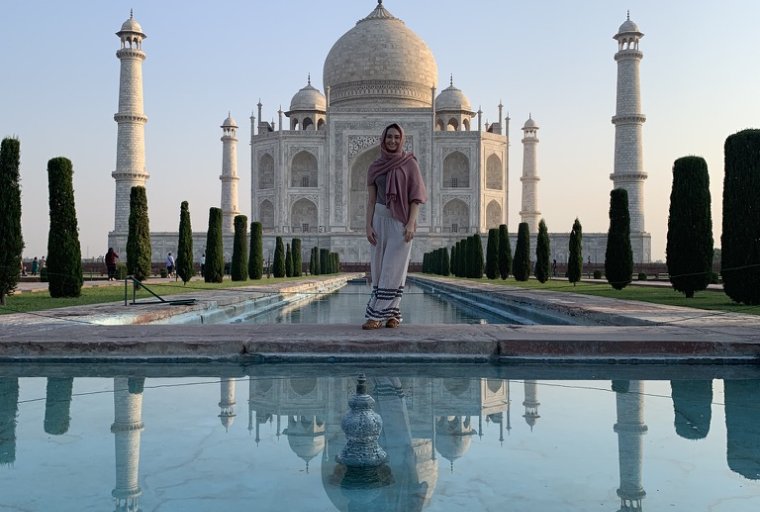 A young woman standing in front of the Taj Mahal with a pink scarf wraped around her shoulders and head