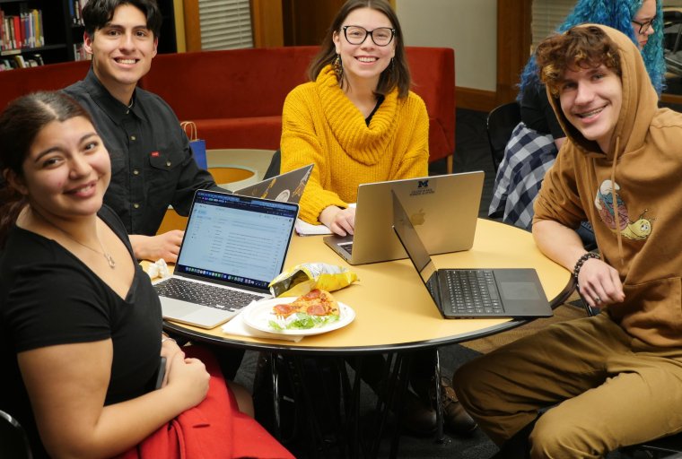 four smiling people sit at a round table and work on laptops