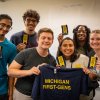 Six people standing, smiling and holding up stickers that say 1st gen. Two people in the center holding t-shirt that say first-gen.