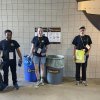 Three stadium volunteers stand in front of bin signage.