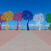 Vibrant metal tree sculptures in red, yellow, blue, and green line a pink walkway under a clear blue sky.
