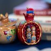 Close-up photograph of two small decorative Armenian objects displayed on a table. On the right is a red, pomegranate-shaped ornament opened to reveal painted figurines of a man and a woman wearing traditional Armenian clothing, framed by small red bead details. On the left is a light wooden pomegranate-shaped container with a removable lid, decorated with hand-painted Armenian motifs in red, green, blue, and yellow. Blurred books are visible in the background, emphasizing the objects as cultural artifacts associated with memory and tradition.