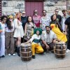 Photo of 14 member interracial troupe posed against an outdoor wall space, with two large drums in foreground.