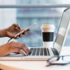 Image of person sitting at a short table holding their phone and working on their laptop, with a coffee nearby.