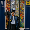 A joyful graduate in University of Michigan cap and gown poses with one arm raised, set against a vibrant mosaic backdrop. Two university banners flank her.