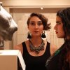 Two women in a museum gallery space; Nabra Nelson stares directly at the camera, while Sarah Fahmy, in profile, views a stone sculpture of a lion’s head.