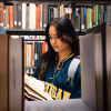 A woman wearing a Michigan sweatshirt reads a book in a library aisle, surrounded by shelves filled with books.