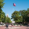 People walking across the Diag with an American flag and Burton Tower in the background, surrounded by green trees under a clear blue sky.