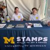 Two students, smiling, sit at a table covered with a blue table adorned with an M STAMPS logo
