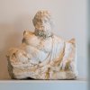White travertine sculpture of the torso and head of Nilus, a bearded and curly-haired male figure in a semi-reclined pose.