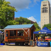A wooden farm stand trailer sits in front of the belltower on S Ingalls mall with shelves filled with local produce and two tents on either side