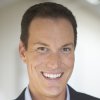 Headshot of Shawn Achor smiling, wearing a dark suit jacket over a light-colored collared shirt against a soft, out-of-focus background.