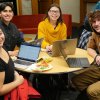 four smiling people sit at a round table and work on laptops