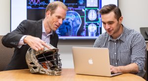 Steve sitting with student at a table studying a laptop next to a lacrosse helmet.