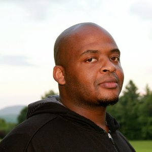 Author Kiese Laymon, an African American man with a shaved head wearing a black zippered shirt.