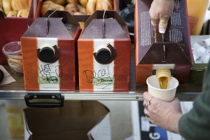 Three Panera box shaped coffee jugs in a row labled left to right, hot h2o, decaf, and coffee. A hand is pictured pouring coffee from the right most jug into a coffee cup that the person is holding. The jugs are on a table with bagels in baskets in the background. Three Panera box shaped coffee jugs in a row labled left to right, hot h2o, decaf, and coffee. A hand is pictured pouring coffee from the right most jug into a coffee cup that the person is holding. The jugs are on a table with bagels in baskets in the background.