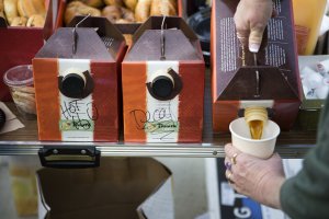 Three Panera box shaped coffee jugs in a row labled left to right, hot h2o, decaf, and coffee. A hand is pictured pouring coffee from the right most jug into a coffee cup that the person is holding. The jugs are on a table with bagels in baskets in the background.