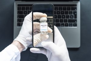 COVID vaccine vial held in gloved hands, seen on the screen of a smartphone above a laptop
