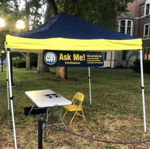 A tent, table, and chair with a sign reading "Ask Me! Info Stations"