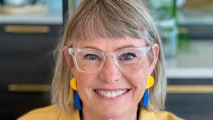 An image of Jules Pieri smiling and looking at the camera. Jules has shoulder-length hair with bangs and is wearing glasses with clear frames. Jules is also wearing maize and blue dangling earrings with a yellow jacket.