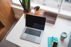 work from home desk setup with a laptop and colorful book, pens, coffee cup, box and plant that overlooks a street with a lot of foot traffic