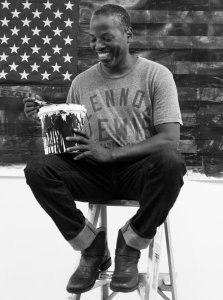 A man sits on a stool with a bucket of white paint, in front of the American flag.