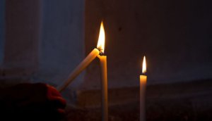 Two candles in a dark room, a hand is lighting a third candle from the first candle.