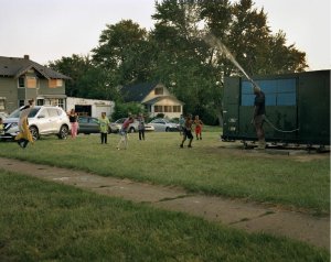 Photograph of a group of people  in a grassy lot playing in water that is being sprayed from a large truck with solar panels