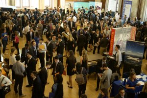 A room full of students who are dressed in business / business casual attire. The students are in lines to speak with various organizations as part of a career fair.